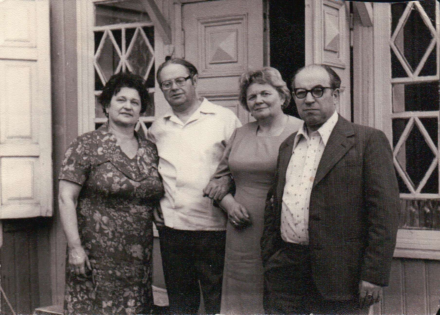 Masha and Alexander with another couple in front of a wooden house with carved window frames.