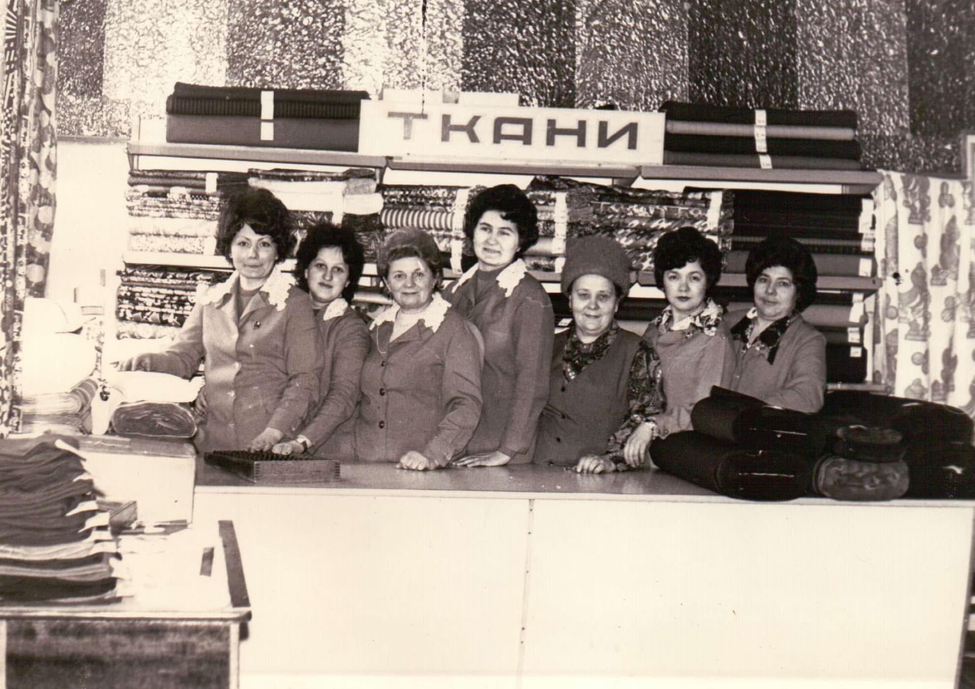 Masha at work behind a counter with coworkers. Sign reads ТКАНИ (Fabrics).