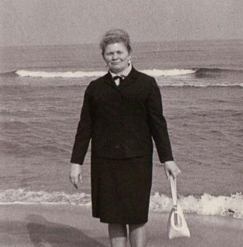 Masha standing alone at the seashore in a dark suit.