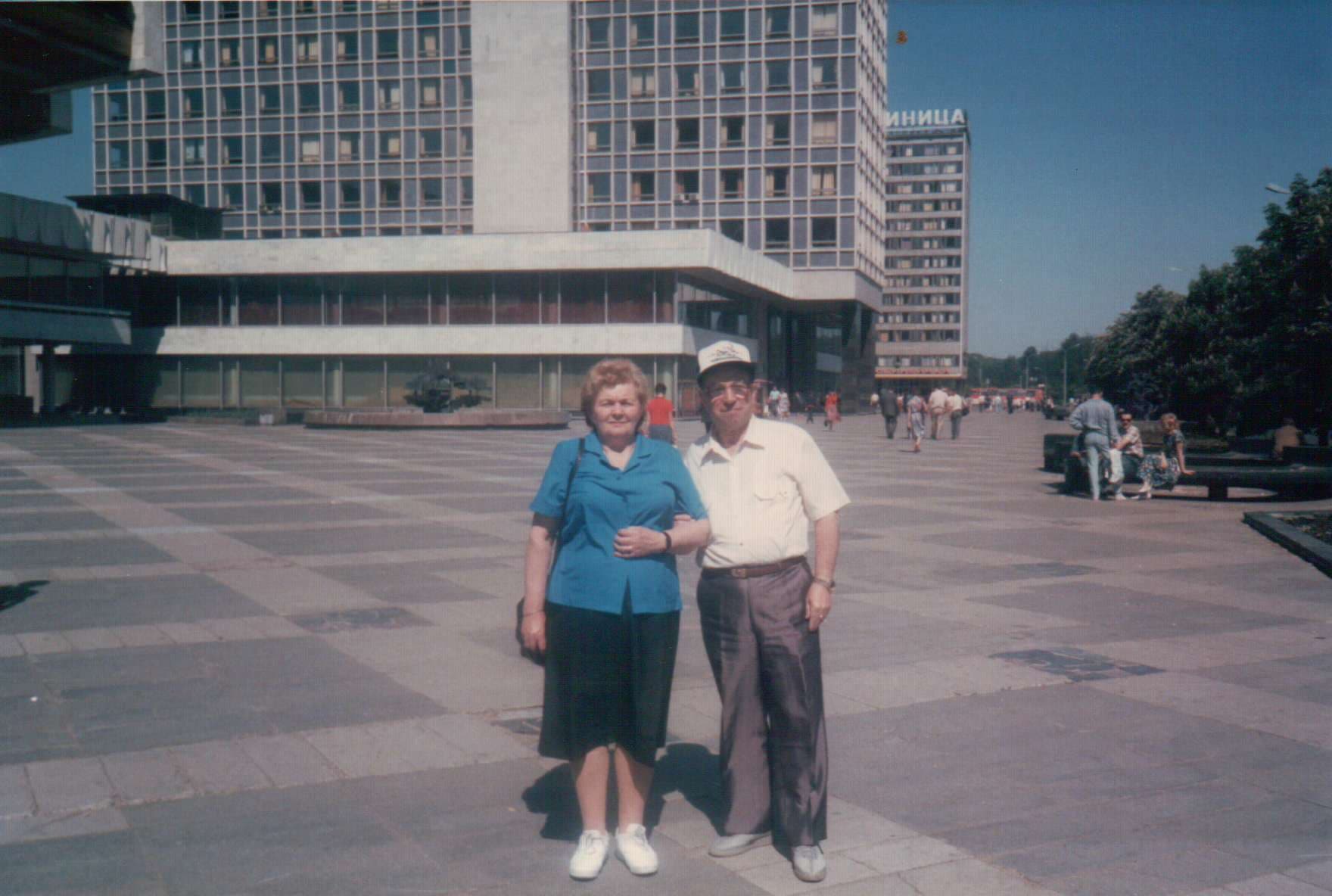 Masha and Alexander in a Soviet city plaza with a modernist hotel building behind them.